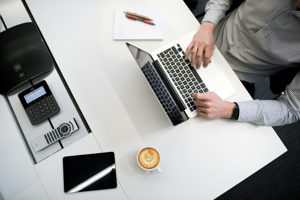 a man seated at a desk using a laptop, with documents and a cup of coffee